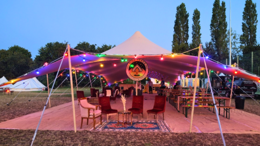 Stretch-tent bar and lounge at the Nine Yards Bell Tents Henley Regatta glamping village, with couches, low tables and festoon lighting in the evening.